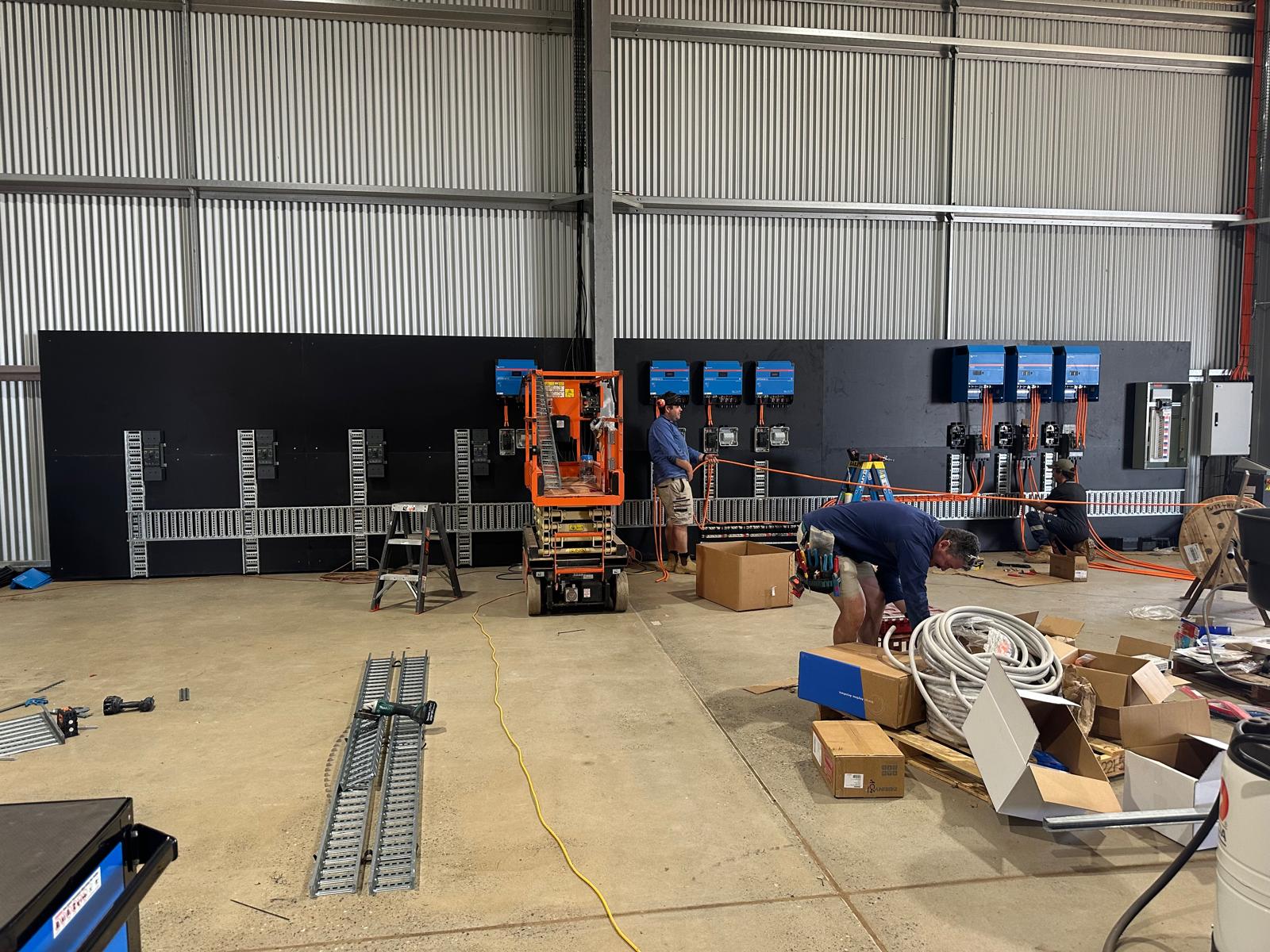 The installation crew wiring Victron equipment inside the farm workshop, surrounded by orange cabling, tools, ladders and a scissor lift.