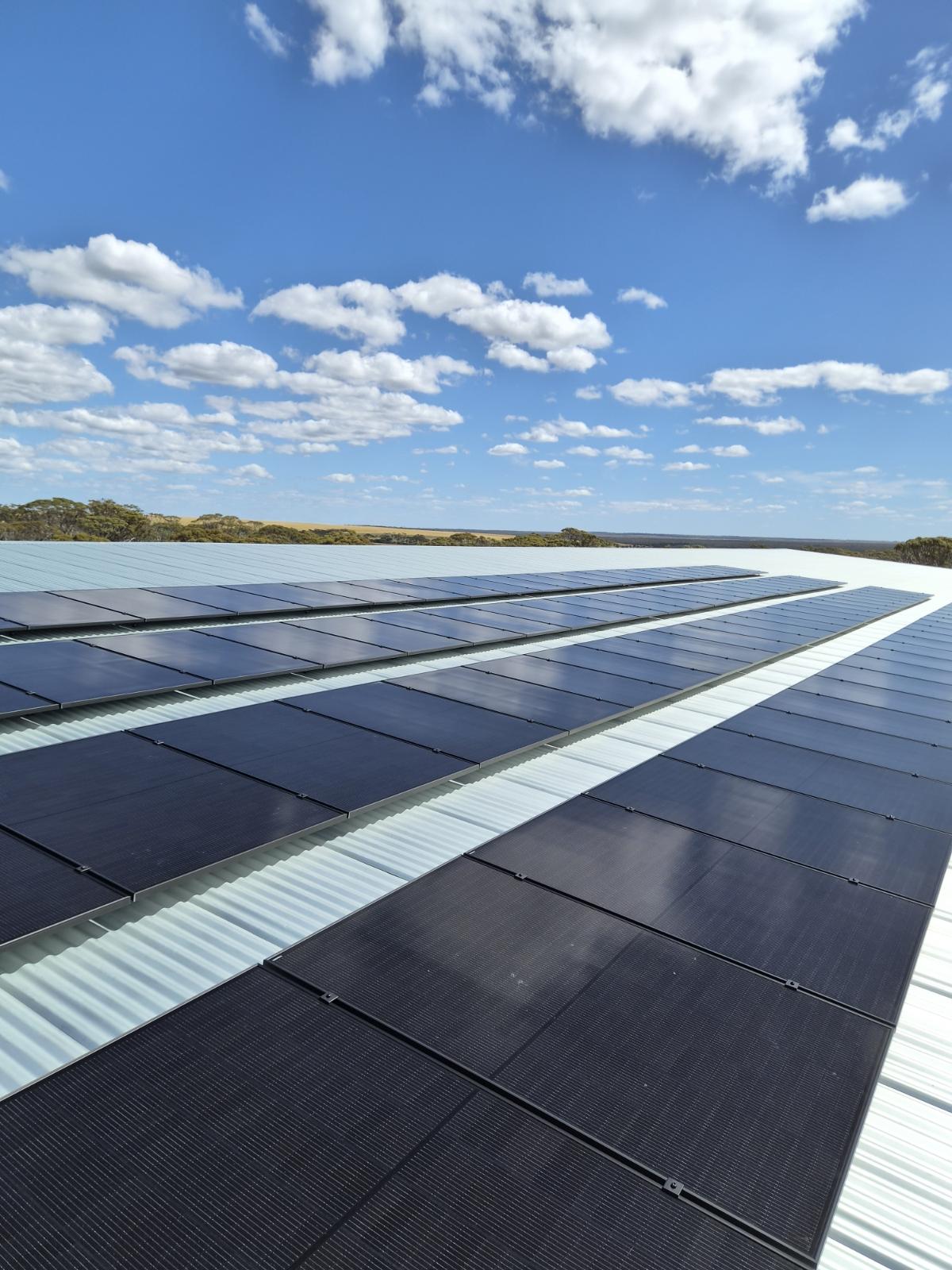 Rows of solar panels across the Phillips River Grazing workshop roof in Ravensthorpe, with farmland stretching into the distance beneath a blue, cloud-dotted sky.